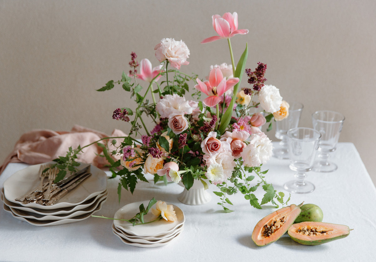 Table setting with floral arrangement, cut melon, and glasses on a white tablecloth.